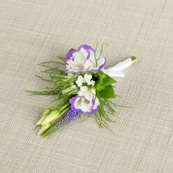 A refined floral boutonniere rests on a pale, finely woven beige fabric, the subtle texture of the material evoking linen laid out on a dressing table before a special occasion in Stanmore. The arrangement is centred around two striking lisianthus flowers, their petals a luminous white with lavishly ruffled edges brushed in vivid purple, creating a clear, elegant contrast that draws the eye. Close beside them, tiny white filler blossoms form delicate clusters, like a soft veil of baby's breath, giving the composition a light, airy feel. Rising through the centre is a slender wand of rich, deep purple muscari, or grape hyacinth, its small, bell-shaped florets stacked tightly together, adding both vertical movement and a deeper note of colour. Fine, feathery greenery, reminiscent of asparagus fern, spills gently around the flowers, its thread-like fronds softening the edges, while a few darker green leaves add structure and a natural, garden-fresh look. Two pale green buds, still closed, peek from the design, suggesting flowers that will open later, much like the quiet anticipation before a ceremony at a local Stanmore church or a reception near Bentley Priory. The stems are carefully aligned and bound with a smooth white satin ribbon, wrapped with precision and finished in a neat bow that signals expert floristry. The overall impression is of calm, modern elegance-an accessory that would sit beautifully on the lapel of a groom, groomsman, or prom guest, its purple-and-white palette echoing twilight skies over Stanmore while keeping the look timeless and understated.