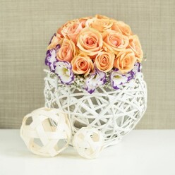 On a pristine white tabletop, a beautifully balanced floral arrangement draws the eye, much like a carefully styled centerpiece at a Stanmore wedding breakfast or engagement celebration. The main cluster forms a near-perfect sphere of peach and apricot roses, with hints of salmon and blush weaving through the palette. Each rose is fully open, revealing intricate spirals of soft petals that press gently against one another, creating a lush, cloud-like dome. Around the base of this rose globe, frilled lisianthus blooms form a graceful collar. Their petals are luminous white with edges dipped in deep violet-purple, the contrast as striking as twilight shadows against a pale sky. Between these flowers, sprigs of white baby's breath thread delicately, their tiny blossoms scattering points of light and adding a weightless, airy dimension. All of the blooms are set into a bulb-shaped white woven container, crafted from interlacing strands of wicker or rattan. Its open, lattice-style construction allows the background to peek through, and the structure casts gentle, crisscrossed shadows that enhance its beautifully handcrafted appearance. This kind of textural detail would sit comfortably in a stylish home off The Common or in a contemporary event space along Stanmore Broadway. In front of the arrangement, resting on the same white surface, two natural-toned decorative spheres made from slender wooden strips echo the basket's weave. One is larger and one smaller, their open, airy patterns adding rhythm and balance to the composition. The backdrop is a softly textured beige fabric that envelops the scene in warmth and calm, highlighting the pastel roses and rich purple accents while creating a mood of intimate, sophisticated celebration.