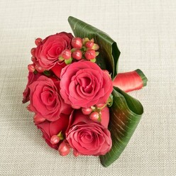 An elegant cluster of deep red roses and berries lies on a pale beige, finely textured fabric, evoking the feeling of a carefully placed bridesmaid bouquet waiting to be carried down the aisle at a Stanmore wedding. The arrangement is compact and rounded, with about six large roses forming a tight, luxurious dome. Each rose displays velvety petals that curl outward from a darker, almost wine-red centre into brighter crimson and scarlet edges, giving the bouquet depth and dimension under the soft, even lighting. Nestled amongst these blooms are rounded Hypericum-style berries in rich shades of red and reddish-pink, their glossy, bead-like forms adding a sense of playfulness and youthful energy, like laughter shared on the way to a ceremony at a local church or venue off Marsh Lane or near Stanmore Country Park. Around the outside, broad, dark green leaves with visible veins are smoothly wrapped and gently folded, creating a neat, contemporary collar that cradles the flowers and gives the bouquet a polished, modern edge. A smooth red satin ribbon secures the stems, its subtle shine catching the light and hinting at the care that has gone into the hand-tying. The neutral, cream-toned background, with its soft fabric texture, keeps the focus firmly on the vivid reds and lush greens, allowing you to almost imagine the faint, green, earthy scent of the leaves and the cool feel of the rose petals. This arrangement feels perfect for bridesmaids, anniversary photographs, or romantic celebrations in and around Stanmore, from homes near Canons Park to intimate garden parties off London Road, where thoughtful, handcrafted flowers help mark meaningful moments.