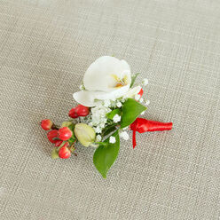 An artfully composed boutonniere is captured from above, lying on a neutral oatmeal-toned fabric that resembles finely woven burlap or linen, its subtle texture creating a calm, natural backdrop. At the centre of the piece is a flawless white Phalaenopsis orchid, its broad petals unfurling like smooth porcelain and curving elegantly away from a softly glowing centre of creamy yellow and gentle orange. Around this focal bloom, sprays of dainty white baby's breath form a light, cloudlike surround, each tiny flower head adding softness and visual movement, as though a delicate mist has settled around the orchid. Beside the white flowers, a striking cluster of round, glossy red berries offers an immediate burst of colour, their surfaces gleaming under the light, with a few pale green, not-yet-ripened buds hinting at natural variety and freshness. Deep and mid-green leaves, some broad and pointed, others more slender, weave through the arrangement, giving it structure and a sense of lushness that recalls the greenery lining the paths around Stanmore Country Park. The stems of the flowers and berries are tightly gathered and wrapped in a vivid red satin ribbon, folded and tied with the kind of precision that comes from many years of floristry practice, leaving short tails that rest neatly against the fabric. This small yet expressive floral design looks ready to be pinned on a groom's lapel for a wedding at a local Stanmore venue, worn at a winter prom, or chosen for a formal holiday gathering, its mix of ivory petals, ruby berries, and rich greenery suggesting both romance and seasonal celebration without overpowering the wearer.