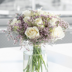 This photograph presents a tender, thoughtfully crafted sympathy arrangement, as if it were resting on a polished sideboard in a Stanmore home overlooking the greenery near Bentley Priory. The main focus is a cluster of pristine white roses, each flower fully open and generously petalled. Their spiralling centres shift from a faintly buttery ivory to a clean, soft white at the outer edges, giving a sense of depth and delicacy. The roses sit closely together but not crowded, allowing each bloom to be appreciated while contributing to the fullness of the bouquet. Lacing between the roses are light, airy sprays of lavender and lilac-pink filler flowers, probably statice, with many tiny, papery blossoms that lend a gentle, mist-like texture. This pastel haze softens the composition and brings a whisper of colour that remains respectfully understated, perfectly suited to moments of remembrance. The flowers are gathered in a tall, slender cylindrical vase made of clear glass, filled with fresh, transparent water. The stems, visible through the sides, are neatly arranged and trimmed to similar lengths, their vivid green providing a natural counterpoint to the pale blooms above. The vase stands on a plain, bright white surface that reflects some of the soft daylight streaming in, enhancing the airy, calm atmosphere. Behind the arrangement, the background is softly blurred into pale neutrals, suggesting a quiet indoor space somewhere near Stanmore station or along The Broadway, where light falls gently and nothing distracts from the quiet beauty of the flowers. Altogether, the image conveys a sense of gentle support and genuine compassion, a fitting floral expression of sympathy crafted with care for the local Stanmore community.