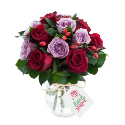 This intimate bouquet pairs saturated red roses with soft lavender companions, arranged in a compact, round silhouette that feels both opulent and serenely composed. Each crimson and burgundy rose opens fully, their layered petals forming plush cups that contrast beautifully with the pale, dusty purple of the lilac roses, producing a visual rhythm that flows from warm to cool. Interwoven throughout are rich, dark-green leaves-likely Salal or Italian Ruscus-whose glossy texture grounds the composition and offers a subtle woodland scent beneath the roses' sweeter notes. Small clusters of bright red-orange rose hips are tucked casually among the stems, their cheerful, tactile presence bringing a touch of country charm and seasonal warmth to an otherwise refined arrangement. The flowers are presented in a rounded, mercury-effect glass vase with an antique silvered finish that shimmers softly under studio light, while a length of natural twine or raffia tied at the neck lends a rustic atelier feel. A white rectangular tag hangs forward showing a stylised rose motif and handwritten-style script in green, signalling a thoughtful gift. Set against a brilliant white background, the colours and glossy reflections read clearly-perfect for an anniversary, a thank-you, or simply to make someone's day. As a local Stanmore florist might suggest, this arrangement works beautifully for delivering a touch of timeless elegance to homes around Stanmore Tube, Stanmore Park, or nearby Harrow.