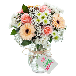 Envision a gentle Stanmore morning, sunlight slipping through lace curtains to illuminate a floral display as thoughtfully composed as a cherished memory. Centered within the arrangement, several peach-pink roses open tenderly, their petals soft and velvety, exuding both warmth and nostalgic romance. Two prominent gerbera daisies in mellow apricot hues with dark, expressive centers bring an immediate sense of optimism, their flawless symmetry reflecting the careful craft honed at Flowers Stanmore's workshop on Church Road. Soft clouds of baby's breath interlace with the other blooms, airy and light, while crisp white chrysanthemums, each showcasing a lime green heart, enhance both freshness and contemporary charm. Occasional sprigs of cool green foliage add movement and hint at walks through Stanmore Country Park. The entire bouquet sits within a clear, gently textured glass vase, the stems slick and vibrant in the water's clarity. Around the vase's neck, a rustic raffia tie offers a tactile finishing touch, from which hangs a rectangular white tag just waiting for a special dedication. With every detail, this bouquet reflects Stanmore's ethos of quality, thoughtful gifting, and the seamless artistry of local flower delivery.