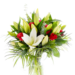 In this carefully arranged bouquet, the eye is led to a single, fully open creamy-white Oriental lily placed slightly forward in a clear, cylindrical glass vase, the pale yellow stamens and gentle green pistil visible like a quiet focal point. Encircling that immaculate bloom, numerous lime-green and soft yellow lily buds promise more blossoms to come, their tight forms adding volume and hope to the design. Interlaced with the lilies are deep, velvety red rosebuds-small, intense pockets of colour whose curled petals speak of affection and timeless romance when set against the lighter tones. The greenery is generous and considered: broad, glossy dark green leaves create depth and support; cascading, long-bladed palm fronds in a lively emerald sway outward to introduce texture and motion; and rounded, silvery eucalyptus stems bring a cool, tactile contrast. The transparent vase reveals crisp green stems in clear water beneath studio lighting that brightens the whites and enriches the reds, giving the bouquet a polished, fresh appearance. Speaking as a local florist in Stanmore, I imagine this arrangement arriving gracefully to homes near Stanmore Station, Canons Park or the High Street - an elegant choice for birthdays, anniversaries or a heartfelt congratulations. Packaged with gentle care, the composition balances purity and passion, scent and structure, and offers both a visual focal point and a fragrant presence that will lift a room with calm celebration. This is the sort of bouquet our customers ask for when they want classic beauty with a modern, airy feel.
