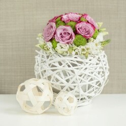 A sophisticated floral centerpiece is captured in a calm, studio-like setting, evoking the atmosphere of an intimate wedding or anniversary dinner in Stanmore. At its base sits a spherical vase crafted from slim white twigs or rattan branches, woven into a loose, open structure reminiscent of a decorative lantern. The intersecting lines form an airy cage that cradles the stems while giving the design a modern yet organic personality. Above this base, the flowers swell into a compact dome, dominated by roses in shades of soft pink and pale lavender. Their petals unfurl in layered spirals, and a few edges deepen into a richer pink, introducing subtle depth and romance. Threaded among the roses are bright green button chrysanthemums, their small, rounded blooms densely packed with tiny petals that add texture and a refreshing burst of colour. Near the rim of the vase, clusters of fine white filler flowers peek out and gently spill over, creating a soft, cloudlike fringe that lightens the composition. The arrangement rests on a clear white surface, and to the left, two decorative spheres made of pale wicker or light wood sit side by side, one larger than the other. Their open, crisscrossed design mirrors the woven vase and completes the crafted, natural theme. Behind the centerpiece, a softly blurred beige background with a subtle woven texture keeps the focus firmly on the flowers while adding warmth. The calm lighting highlights each petal and strand, making this image ideal for representing a refined wedding table arrangement or elegant home décor piece available from local Stanmore florists who understand both design and detail.