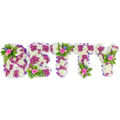 This bespoke memorial piece spells BETTY in solid, freestanding capital letters and is composed with an attention to balance that comfortingly reads as both personal and ceremonial. The primary surface of each letter is densely packed with crisp white chrysanthemums whose rounded heads create a soft, cloud-like texture; these provide a quiet, peaceful field against which deep magenta-purple chrysanthemums form defining inner patterns and outlines, giving the lettering sculptural clarity. A narrow purple ribbon is discreetly tucked along the sides of each letter, unifying the palette. Perched thoughtfully on the B, across the top of the central T, and at the upper arm of the Y are small, natural clusters of flowers: pale pink roses in mixed stages of bloom, tiny bright-blue blooms suggestive of cornflower or delphinium, and fresh fern fronds and greenery that add a garden-like softness and subtle scent. The arrangement is photographed on a pristine white background with gentle studio light that reveals petal texture, shadowed contours, and the layered workmanship. As a local touch, this is the type of custom tribute we craft at Flowers Stanmore for families gathering in Stanmore or at nearby Canons Park and Stanmore Tube, where personalised floristry helps express grief and remembrance. The overall mood is restrained yet affectionate - an elegant, tactile memorial that reads as a final salute, a name held tenderly in flowers and memory.