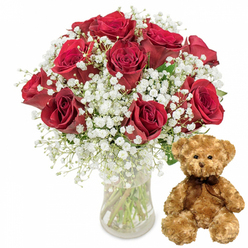 This photograph, composed against a smooth white backdrop, captures a timeless romantic gesture that would not look out of place in a Stanmore living room on a special occasion. On the left side, a clear glass vase holds a generous bouquet of around a dozen deep red roses, each blossom full and velvety, with tightly layered petals that curl gently at the edges. The roses stand at varying heights and stages of openness, combining to form a lush, rounded silhouette that feels rich and abundant. Threaded throughout the bouquet, delicate sprigs of white baby's breath (Gypsophila) add contrast and softness, their tiny white blooms forming light, airy clusters that seem to float between the roses like small cotton clouds. The glass vase is understated yet elegant, allowing the green stems and the freshness of the water to be seen, reinforcing the sense of a professionally arranged gift, perhaps prepared by Flowers Stanmore for same-day delivery. To the right of the floral display, a plush teddy bear in light brown fur sits upright, slightly angled towards the flowers as if sharing in the sentiment. Its fur looks wonderfully soft and gently textured, and its classic details-small dark button eyes, a neat embroidered nose, a subtle stitched smile-give it a gentle, reassuring personality. Around its neck, a dark brown ribbon is tied in a simple bow, adding a finishing touch that makes the bear feel ready to be gifted. Together, the vibrant roses, airy gypsophila, and cuddly bear create an impression of romance and tenderness, perfect for anniversaries, birthdays, or Valentine's surprises in and around Stanmore, from homes near the station to quieter streets leading towards Bentley Priory.