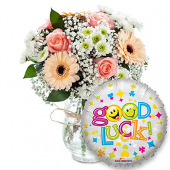 This uplifting Good Luck ensemble reads like a small celebration captured on white studio light, perfect for sending encouraging wishes around Stanmore. A full bouquet sits slightly left of centre in a clear glass vase, the neck wrapped in rustic twine that hints at hand-tied craftsmanship. Soft blush-pink roses and warm peachy-orange Gerbera daisies with their dark brown centres create a romantic, upbeat colour story; interspersed are airy sprays of white baby's breath and compact clusters of white-and-lime chrysanthemum florets that add crisp contrast and a subtle, green-tinged freshness. Deep green foliage peeks through, lending structure and a cool leafy scent beneath the sweeter rose notes. To the right, overlapping the bouquet's lower edge, a shiny silver Mylar balloon declares Good Luck! in buoyant, multicoloured letters; two yellow smiley-face o's-one winking, one sticking its tongue out-bring a playful human touch, while scattered hearts, stars and tiny coloured shapes sparkle across the balloon's surface. I picture this arrangement being chosen for exam send-offs, auditions, or moving-day well wishes, and as a local shop I know Flowers Stanmore often prepares similar sets for same-day delivery across Stanmore station, the High Street and nearby residential roads. The composition balances texture and colour, scent and sentiment, offering a comforting, joyful send-off in one carefully composed gift.