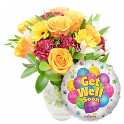 A cheerful, thoughtfully composed "Get Well Soon" arrangement presented on a pristine white backdrop: a generous, hand-styled bouquet in a clear glass vase sits slightly left-of-centre, while a glossy round Mylar balloon overlaps the lower right, declaring an upbeat message. The floral mix is warm and uplifting-plush orange and golden-yellow roses form the focal point, their petals soft and layered, emitting a subtle, citrus-like aroma that feels like sunlight in bloom. Bright yellow gerbera daisies add a playful, sunlit energy, contrasting with the deep maroon carnations that provide depth and a velvety texture. Sprays of white alstroemeria punctuate the palette with delicate points of light, and varied green foliage cradles every bloom to give the bouquet a natural, garden-fresh silhouette. The clear vase reveals clean stems and water, a small sign of the florist's attention to freshness. The balloon's reflective silver surface frames a cluster of cartoon balloons in purple, pink, blue, green and yellow-some with stripes, some polka-dotted-while bold, cheerful yellow-and-blue lettering announces "Get Well Soon." Tiny starbursts edge the design and a discreet "KALEIDOSCOPE" logo sits at the bottom rim. As a vanishingly personal touch from Flowers Stanmore, this arrangement is perfect for lifting spirits-suitable for bedside delivery to the Royal National Orthopaedic Hospital, a table in a Stanmore home, or same-day delivery across Stanmore Broadway and surrounding streets.