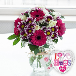 Set against a softly blurred, luminous background reminiscent of a sunlit window in Stanmore, this floral arrangement and balloon pairing feels like a snapshot of pure celebration. A clear glass vase takes centre stage, its rounded shape holding a carefully structured bouquet. Inside, neatly trimmed stems are visible through the water, revealing the craftsmanship behind the design. Rising above the vase, large gerbera daisies in a vivid magenta dominate the scene, their wide, velvety petals radiating from dark, textured centres that ground the composition. Surrounding them, generous clusters of small chrysanthemums create a sense of abundance, each tiny bloom adorned with deep purple petals whose tips fade into crisp white, highlighting cheerful yellow centres that seem to glow. Creamy white roses are artfully tucked throughout the arrangement, their softly cupped petals adding a note of sophistication and tenderness. Delicate sprays of white gypsophila float like a delicate lace over and between the larger blooms, lending a dreamy, romantic softness. Fresh, verdant foliage pushes through at the edges and between flowers, adding depth and the reassuring presence of garden greenery. Around the neck of the vase, rustic twine is tied in a simple bow, suspending a neatly cut white tag decorated with a pink rose graphic and handwriting-style text, adding the warm feel of a personalised gift, as if created especially for someone in Stanmore or nearby Canons Park. Just to the right of the vase, a shiny heart-shaped foil balloon leans into the frame, bordered in reflective silver and filled with bold, multicoloured letters spelling out an affectionate phrase and a playful "24-7," emphasising constant care and connection. Resting together on a pale surface, the flowers and balloon radiate joy, making this scene ideal for marking birthdays, romantic milestones, or heartfelt get-well wishes across the Stanmore area.