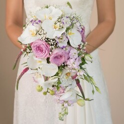 This exquisite, eye-level bridal portrait, taken in Stanmore, reveals a bride gently embracing a luxurious waterfall bouquet-a visual ode to the neighborhood's refined tastes and passion for local blooms. The bouquet flows gracefully from her hands, its composition dominated by pristine white Phalaenopsis orchids, their subtle golden hearts gleaming softly beneath serene light. Around them unfurl plump roses in pale pink and lavender, their lush, velvety petals releasing a hint of sweet fragrance evocative of fresh florals from Canons Park or the local weekly market. Airy clouds of white baby's breath float gently between the grander blooms, adding an ethereal softness. Vibrant magenta amaranthus cascades in long, tactile strands, their unique texture grounding the arrangement. Accents of light lilac blossoms and slender buds weave through, bringing dimension and life, while touches of green foliage hint at Stanmore's open spaces. The bride's white gown, with its subtle lace motifs, frames the bouquet with timeless grace. Together these elements create a harmonious, sophisticated palette and celebrate the subtleties that make Stanmore weddings feel so special. Every detail, from flower choice to the arrangement's elegant flow, reflects the expertise and artistry of the area's talented florists, ensuring the bouquet will be remembered as a symbol of love's promise.