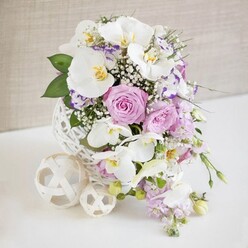 An elaborate teardrop-shaped bouquet is shown from a slightly elevated angle, capturing the way its flowers cascade gracefully downward in a classic bridal waterfall style, perfectly suited to a Stanmore wedding. At the top of the arrangement, blooms gather densely in a lush cluster, then gradually trail toward the lower tip, creating a sense of movement and natural flow. Large white orchids, most likely Phalaenopsis, dominate the composition, their petals smooth and luminous, flaring outward like elegant wings. Each orchid has a delicate center touched with yellow and gold, adding a hint of warmth and luxury. Between and around these orchids, light pink and pale lavender roses nestle in tight groups, their soft, velvety petals spiralling inward, evoking romance and quiet tenderness. Wispy sprays of white baby's breath weave throughout the bouquet, their tiny blossoms forming a fine mist that softens the transitions between flowers and adds an airy, cloud-like quality. Along the descending curve of the bouquet, subtle touches of lilac and muted purple blooms echo the colour of the roses and guide the eye downward, enhancing the waterfall effect. Fresh green foliage peeks out in slender, carefully placed leaves, giving the piece a natural rhythm and a hint of woodland freshness while maintaining the overall pastel palette. The bouquet is supported by a white decorative base that appears textured, perhaps ceramic or lace-inspired, with several smaller white spheres nearby featuring intricate cut-out patterns, reinforcing the bridal, almost ethereal theme. The entire design is set against a simple cream or off-white background, softly lit so every petal, shadow, and curve stands out. It feels like the perfect bouquet for walking down the aisle in Stanmore, whether at a local church, an elegant reception venue, or a private garden celebration, conveying luxury, devotion, and serene beauty.