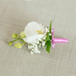 A single, immaculate white orchid boutonniere is captured resting on a light beige, linen-textured fabric, its horizontal placement giving the image a sense of calm balance, like a quiet pause before guests arrive at a Stanmore wedding or black-tie celebration. The orchid, likely a Phalaenopsis, is the clear focal point, with broad, satin-smooth petals that open outwards in a perfect, rounded shape. At its centre, delicate details of soft yellow and the faintest whisper of pale pink add warmth and depth, drawing attention to the intricate heart of the bloom. Surrounding the base of the orchid is a soft halo of white baby's breath, each tiny Gypsophila blossom forming part of a light, cloud-like cluster that lends the design an ethereal, almost dreamy quality. A few slender, bright green leaves emerge from behind the flowers, offering a fresh, natural contrast and evoking the leafy streets and gardens around Stanmore Hill and Canons Park. Off to one side, a short stem bears several tightly furled, lime-green orchid buds, hinting at growth and the promise of future flowering moments, much like a couple just starting their journey together. The stems are carefully gathered and wrapped in a bright fuchsia pink satin ribbon, tied into a neat, refined bow. The ribbon's glossy surface catches the soft, even lighting, adding a joyful pop of colour against the neutral background. The gentle light enhances every texture: the orchid's velvety petals, the feathery baby's breath, and the subtle weave of the linen. This boutonniere feels perfectly suited to a groom, usher, or prom guest attending a special event in Stanmore, offering understated elegance and timeless charm.