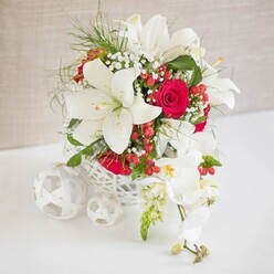 A sophisticated floral tableau is arranged on a flawless white surface, set before a soft beige backdrop that looks like gently gathered fabric, capturing light in the same understated way as a stylish Stanmore boutique venue. The focal point is a round, white basket-shaped container with an intricate open weave, its airy lattice pattern revealing glimpses of stems and adding a contemporary yet handcrafted touch that would sit beautifully on a wedding top table near Canons Park. Rising from this delicate base, several large white oriental lilies open generously, their sculpted petals fanning outward to reveal elegant stamens tipped with warm golden pollen. Intertwined among them, rich red roses-deeply saturated and velvety-remain mostly in tight bud, creating pockets of intense colour that pull the eye through the design. Sprays of white baby's breath drift lightly around the larger blooms, adding tiny, star-like flowers that soften every transition and create an almost misty halo. Clusters of glossy red hypericum berries provide charming flashes of festive brightness, suggesting seasonal celebrations or winter weddings in the Stanmore area. Airy green foliage, slender and feather-light, breaks up the mass of petals, lending the arrangement a sense of breath and natural ease. To the right, a cascade of white phalaenopsis orchids arches gracefully, their broad petals forming delicate, moth-shaped faces with subtle yellow and blush centres, while a few buds at the tip remain smooth and unopened. On the left side of the scene, white woven spheres rest on the tabletop, mirroring the container's texture and anchoring the composition visually. The overall effect is one of refined romance and polished celebration, perfectly suited for wedding receptions, anniversary dinners, or elegant gatherings across Stanmore, from The Broadway to the quiet residential lanes.