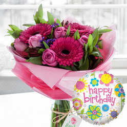 An exuberant birthday arrangement is captured in gentle daylight, evoking the feeling of a surprise delivery arriving at a Stanmore townhouse just off Marsh Lane. Centre stage is a lavish bouquet of fresh flowers set in a clear glass cylinder vase, the stems clearly visible through the water, wrapped at the base with layers of translucent blush-pink cellophane that form a soft, fluttering collar. The bouquet is dominated by striking gerbera daisies in rich fuchsia and magenta tones, their wide, symmetrical petals circling deep, almost black centres that draw the eye. Interlaced with these are clusters of delicate pink roses, ranging from tightly furled buds to half-open blooms, each petal gently curling to reveal a creamy interior, suggesting sweetness and affection. Between the main flowers, tiny clusters of deep purple filler blossoms add texture and a cool contrast, while generous green foliage fans out, giving the design a lush, rounded shape reminiscent of a well-kept Stanmore garden after spring rain. A rustic length of twine is tied around the neck of the vase, holding a small, off-white rectangular gift tag subtly printed with a rose emblem, hinting at a premium, carefully chosen gift rather than something hurriedly bought. In the lower right foreground, almost leaning into the flowers, floats a round foil balloon with a crisp white base, lightly sprinkled with grey. Brightly coloured floral and circular motifs in yellow, sky blue, lime green, pink and purple dance around its edge, framing the central message "happy birthday" written in bold pink cursive. The background is softly out of focus, suggesting an airy indoor space, which helps the vivid blooms and playful balloon stand out as a joyful, ready-to-send birthday surprise from a trusted local florist.