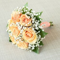 Resting atop a subtly textured, pale linen and bathed in soft natural light, this bouquet is a symphony of peach-hued roses and delicate white baby's breath, assembled with an artist's touch befitting Stanmore's refined floral heritage. Each rose, opening from a tight central spiral to broad, ruffled petals, blushes with shades of apricot and soft orange, evoking the first light over Canons Park. The dreamy cloud of baby's breath woven throughout provides visual lift and softness, cradling the roses in airy contrast and imbuing the arrangement with an almost otherworldly grace. Deep green leaves punctuate the composition, their sheen and depth reminiscent of Stanmore's private gardens, adding support and verdant richness. The handle is carefully wrapped with a coral-peach satin ribbon, cool and smooth, its surface spiralling with hand-placed, glossy faux pearls-a bridal embellishment that shimmers in the filtered daylight. The effect is nothing less than sophisticated, tactile luxury: the bouquet feels full-bodied yet light, like an embrace ready for a bride crossing the lawns of Bentley Priory. Every petal's nuance and every pearl's lustre are gently revealed by the lighting, making this bouquet the perfect companion for weddings, engagements, or anniversary celebrations in Stanmore and beyond-a floral design to linger in memory, exuding grace and joy at every glance.