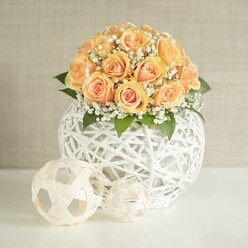 An artfully composed floral display features a generous dome of peach-toned roses arranged with the precision and care of an experienced Stanmore florist. Around twenty to thirty roses are clustered so tightly their overlapping petals form a continuous cushion of soft apricot and warm orange, with deeper, glowing centres that fade to lighter, velvety edges. Tiny white blooms of baby's breath are tucked in between, their frothy, cloud-like texture giving the arrangement a sense of lightness, as if the flowers were resting on mist. Occasional dark green leaves peek through, offering contrast and reminding you these are fresh, living stems, much like those selected each morning from markets serving north-west London. The bouquet is nestled into a striking spherical container made from interlaced white strips, woven into an airy lattice orb that is both modern and gently rustic, reminiscent of contemporary interiors along Stanmore Hill. On the crisp white surface beneath, two smaller woven balls sit just in front of the vase, one slightly larger than the other, in creamy beige tones that echo the texture without competing with the flowers. The background is a soft, neutral wall in pale beige or greige, with a faint linear texture that catches the light without drawing attention. Everything is lit with calm, even daylight, casting delicate shadows and highlighting each curve of petal and strand of wicker. The scene evokes a sense of quiet celebration, ideal as a centrepiece for a wedding breakfast in a Stanmore venue, a refined birthday table, or a thoughtfully styled home just off The Broadway.