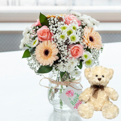 A tender anniversary arrangement is displayed on a pure white surface, glowing in soft, bright light that feels reminiscent of a sunny room in Stanmore. At the heart of the image is a clear glass jar-style vase brimming with a carefully balanced bouquet. Blush pink roses, their petals unfolding in velvety spirals, mingle with peach-hued gerbera daisies, each bloom defined by its rich dark brown centre. Together they form the warm core of the design. Encircling them are clusters of crisp white chrysanthemums, some tipped with fresh green, and airy sprigs of white baby's breath that weave delicately between the larger flowers, giving the piece a romantic, cloud-like softness. The stems are neatly gathered and visible through the transparent glass, immersed in clear water that speaks to the bouquet's freshness. Around the neck of the vase, a piece of natural twine is tied in a casual bow, adding a rustic note. From this bow hangs a small white rectangular tag, bearing a simple pink heart and elegant green handwriting that suggests a handmade or personalised message, as you might find with a bespoke order from a Stanmore florist.   

To the right, a cream plush teddy bear sits upright beside the vase, slightly angled towards the blooms. Its fur appears fluffy and inviting to the touch, with small, dark, bead-like eyes and a brown button nose that give it a gentle, comforting expression. A narrow dark brown satin ribbon is knotted neatly around its neck, echoing the deeper tones in the flowers. The background is softly blurred and flooded with natural light, creating a serene atmosphere that frames the gift set beautifully. This scene captures the emotion of an anniversary celebration in Stanmore-calm, thoughtful, and full of quiet affection, well-suited to a surprise delivery to homes near Stanmore Hill or a cosy evening in with someone special.