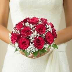 Just steps from Stanmore's stately homes and tranquil parks, a bride's hands gently enfold a bouquet that radiates heartfelt tradition and grace. The arrangement is a masterful blend of classic wedding blooms: rich, velvety red roses are nestled tightly together at the centre, each bud and blossom exuding the passion and elegance of enduring love. Tucked between the roses, delicate clusters of baby's breath add a soft, cloud-like frame, their miniature white flowers casting a spell of enchantment over the bouquet. Lush green foliage peeks from beneath, invoking memories of late spring walks along Stanmore Hill. The bride's manicured fingers wrap securely around the stems, and her wedding gown's intricately patterned lace or embroidery forms a beautiful backdrop. A smooth, flowing sash trails from the bodice, enhancing the gown's fluid lines and romantic feel. Gentle, diffused light creates a warm and inviting atmosphere, while a blurred background in subtle beige tones ensures every detail of the bouquet is illuminated. Prepared locally by Flowers Stanmore, this arrangement is ideal for those seeking inspiration for classic, meaningful bridal florals in North West London.