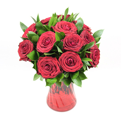 An opulent cluster of approximately twelve to fifteen deep red roses is arranged in a classic round formation, resting in a translucent red glass vase and set against a pure white background that lets the colours take centre stage. Each rose is fully opened to its most flattering point, revealing intricate spirals of velvety petals that layer tightly at the heart and soften towards the edges, giving the blooms a plush, almost satin-like appearance. The red is a rich, ruby shade with subtle variations where light brushes across the petal curves, suggesting freshness and careful selection by a skilled florist. Threaded throughout the bouquet is a substantial amount of dark, glossy green foliage, likely ruscus or similar sturdy greenery, its smooth leaves peeking out confidently between the blooms, adding rhythm and depth to the arrangement. The foliage shapes the bouquet into a full, rounded silhouette while ensuring the roses remain the star. The red glass vase, with its gently flared lip and softly tapered body, has a delicate ribbed or waved texture that catches reflections, so the vase itself echoes the movement of the petals above. Because the glass is semi-transparent, there are hints of stems and layered tones of garnet and cherry red visible through it. Lit with even, neutral lighting, the bouquet casts only subtle shadows, keeping the overall mood clean, modern, and serene. This image feels like a romantic gesture waiting to be carried along Marsh Lane or into a restaurant near Stanmore Broadway for a special dinner, a floral composition that speaks clearly of love, admiration, and celebratory moments shared in this corner of north-west London.
