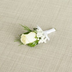 This image features an elegant, handcrafted boutonniere placed at a slight angle on a pale beige, linen-like fabric that feels very much at home in a refined Stanmore wedding setting. At its centre is a single ivory white rosebud, its petals still partially closed, smooth and velvety, with a gentle curl at the edges that hints at the bloom it will soon become. A soft wash of light glides over the rose, picking out the layered structure of each petal and giving the flower a subtle glow, as though it has just been selected from a fresh delivery destined for a ceremony near Stanmore Country Park or Bentley Priory. Surrounding the rose is a delicate halo of white baby's breath, each tiny blossom like a snowflake, adding airy texture and soft contrast against the central bloom. Complementing these are slender, vibrant green leaves and a few fern-like fronds that arch and tilt around the flower, framing it with natural movement and colour. The stems are tightly gathered and wrapped in a smooth white satin ribbon, the finish precise and clean. A small, perfectly tied bow sits just below the flower heads, signalling the meticulous craftsmanship of a professional florist. The background fabric's fine weave and understated tone create a neutral stage, allowing the boutonniere's whites and greens to stand out without distraction. The overall mood is calm, romantic, and contemplative, ideal for a groom, best man, or usher walking through the leafy streets of Stanmore towards a waiting wedding car. The image conveys the quiet joy, expectation, and formal charm that surround a carefully planned wedding day.