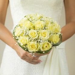 Bathed in the gentle glow of a Stanmore afternoon, this bouquet embodies understated romance and refined charm. Creamy yellow roses spiral from the heart of the arrangement, each one a luminous testament to expert floristry inspired by walks through Stanmore Country Park. Sprigs of baby's breath, light and feathery, are nestled amidst the roses, drifting like the first trace of dawn on a cloudless day, while bands of sleek, dark green foliage coil gracefully at the base, each curve a nod to the elegant grounds of the local manor houses. The bride's hands, neat and softly blushed at the nails, hold the bouquet with loving care, a simple ring on her finger catching the ambient light and echoing the spirit of joyful vows. In the background, a subtly textured white wedding gown recedes into a gentle blur, ensuring the bouquet's colours and textures remain the focus for both memory and admiration. The composition breathes with life and tradition, a celebration of Stanmore's rich landscape and the enduring beauty found in every wedding bouquet expertly arranged for ceremonies in this welcoming neighbourhood.