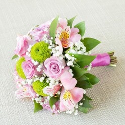 A lush, horizontally placed bouquet stretches across a pale, finely textured fabric, as though set down carefully on a linen-covered table in a Stanmore townhouse just off The Broadway. Soft pink and blush-lilac roses form the romantic core, their petals spiralling inwards with velvety precision, some blooms still tightly formed while others open generously to reveal layered hearts. Between them, bright lime-green button chrysanthemums provide a cheerful spark, their compact, rounded forms and crisp petals giving the piece a lively, tactile energy. Light pink alstroemeria thread through the roses and chrysanthemums, their slim, flared petals revealing soft yellow throats streaked with fine maroon speckles, like delicate brushstrokes. Tiny tufts of white gypsophila hover between the larger flowers, adding gossamer lightness and making the bouquet feel cloudlike and full without being heavy. The blossoms are cradled by rich green foliage that frames the arrangement, some leaves offering broad, glossy surfaces, others narrower and more textured, lending structure and depth. At one end, the exposed stems are neatly aligned and tightly wrapped in a satin ribbon of vivid magenta or fuchsia, its smooth, slightly reflective finish giving the bouquet a polished, gift-ready look while leaving the natural stem ends charmingly visible. The gentle, even lighting highlights the freshness of each petal and the subtle weave of the background fabric, suggesting careful, professional floristry. The overall effect is that of a springlike, joyful bouquet, ideal for a bridesmaid walking down the aisle in Stanmore, a milestone birthday, or a same-day delivery surprise that brings fragrance, colour, and a sense of cherished occasion into a local home.