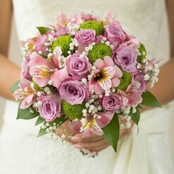 Framed by the intricate lace of a wedding gown and the genteel charm of Stanmore's conservation areas, this lush round bridal bouquet bursts with detail and colour. Soft lavender and blush pink roses unfold at its core, their petals layered in cool, romantic tones that exude a gentle fragrance with every fresh bloom. Intertwined among them, crisp green button chrysanthemums supply a lively, spherical texture, sparking playful energy reminiscent of the vibrant flowerbeds found near Stanmore Recreation Ground. The pink alstroemeria, with their delicate maroon-streaked petals, add both visual and aromatic richness, while wisps of white baby's breath create a soft haze, knitting together each element into a harmonious whole. Deep, glossy green foliage provides a natural underpinning, enhancing both depth and structure. Meticulously arranged by a Stanmore florist, this bouquet feels both deeply personal and artfully designed, compellingly suited for elegant wedding celebrations or heartfelt vow renewals around Stanmore and neighbouring villages. Every nuance combines to evoke the spirit of new beginnings, cherished traditions, and the beauty of local craftsmanship.