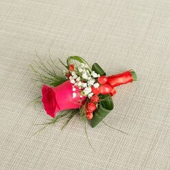 Displayed horizontally on a pale beige, gently woven fabric, this handcrafted boutonniere appears as if it has just been set down on a suit jacket waiting in a Stanmore dressing room before a ceremony. The eye is drawn first to a luxurious fuchsia-pink rose, its petals tightly furled at the centre and gradually loosening into a soft spiral of velvety layers, suggesting both tenderness and confident colour. Surrounding the rose's base is a soft collar of white baby's breath, the tiny blooms like a cloud of lace that brightens the composition and adds a sense of purity. Tucked between these white clusters are plump, shiny red hypericum berries, their spherical shapes and saturated hue giving a subtle, joyful energy, reminiscent of festive gatherings around Stanmore Hill or family photos in Canons Park. The greenery has been arranged with a florist's practiced hand: slender, feathery sprigs of fern-like foliage stretch gently outward, while darker, broader green leaves are folded and curled into a supportive cradle that frames the rose and keeps the design compact. At the bottom, the stems are bound together with a rich red satin ribbon, wrapped smoothly and tied into a neat, understated bow that mirrors the rose's tone and adds a touch of luxury you can almost feel beneath your fingers. Soft, even lighting from above casts delicate shadows across the textured background, accentuating the contours of each petal and leaf. The overall mood is romantic and composed, perfect for a groom, groomsman, or formal guest having flowers delivered locally from Flowers Stanmore for a heartfelt celebration.