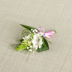 On a pale, linen-like background that feels as soft and neutral as a freshly pressed suit jacket, a compact floral boutonniere is laid out with the care of an experienced Stanmore florist preparing for a wedding morning. The central bloom is a white alstroemeria, its petals slightly flared and gently ruffled at the tips, creating a cup that reveals subtle reddish-brown speckles near the throat. These delicate markings add character to the blossom, drawing attention to the stamens nestled inside and giving a hint of natural wildness to an otherwise polished flower. Beside it, on the left, rises an elongated cone of tightly packed lime green buds, almost architectural in form, each small bud forming part of a textured spike that feels both contemporary and fresh, reminiscent of new shoots seen in the parks and leafy streets around Stanmore Hill. The arrangement is softened by clusters of cloud-like white baby's breath, their tiny, starry flowers hovering between the larger elements and creating a veil of lightness. A base of deep green foliage underpins everything: broad, dark leaves spread like a quiet backdrop, while fine, grass-like strands curl and arc, lending a sense of motion and natural ease, as though caught in a gentle breeze. At the right, a sleek pink satin ribbon gathers the stems into a tidy bundle, the bow small but precise, adding a pop of upbeat colour that hints at romance and celebration. Even, diffused lighting reveals the fresh textures of every petal and leaf, suggesting that this boutonniere is newly made and ready for same-day collection or local delivery around Stanmore, ideal for groomsmen, prom dates, or anyone dressing up for a special occasion.