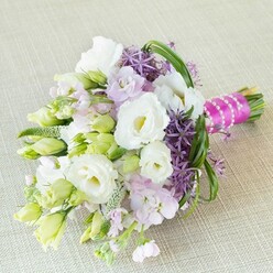Bathed in even natural light atop a subtly textured cream fabric, this hand-crafted bouquet by Stanmore's florists is a symphony of spring celebration. Central to the arrangement, pristine white Lisianthus-petals gently ruffled as if stirred by the morning breeze in Canons Park-open beside slender green-tinged buds, symbolising both hope and tradition. Around them, clusters of soft lavender-pink stock infuse the air with delicate, fleeting perfume, their gentle colouring mirroring the blush of first love and friendly laughter that resonate through Stanmore's wedding venues. Bold, globe-like allium flowers in deep purple, with their starburst florets, stand poised amidst the pastels, introducing architectural intrigue and a sense of playful rhythm. Every space is filled with texture-lime-green buds, glossy filler sprigs, and the looping movement of bright bear grass create a tapestry of verdant freshness, recalling walks through Stanmore's shaded lanes. The stems are tightly gathered and secured with a radiant fuchsia ribbon, finished by a winding, luminous string of pearls that nods to classic bridal details and contemporary flair. This bouquet is more than a visual delight; it encapsulates the energy, unity, and sense of occasion that mark unforgettable weddings and celebrations in the heart of Stanmore.