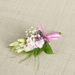 A small but exquisitely detailed floral corsage or boutonniere is displayed on a pale beige, finely woven linen cloth, the fabric's gentle texture adding a calm, neutral backdrop that lets the flowers truly breathe. In the centre, a tight gathering of soft pink, multi-petalled blooms unfurls with a ruffled, almost cloud-like effect, the petals moving from a slightly deeper blush at their base to a whisper-light tint at the edges, like a Stanmore sunrise seen over leafy back gardens. Threaded delicately through this pastel heart are numerous sprigs of classic white baby's breath, their minuscule blossoms forming a lace-like haze that softens and lightens the whole arrangement. Touches of lavender and deeper purple from tiny spiked florets introduce a quiet sophistication, grounding the sweetness of the pinks and whites without disturbing the boutonniere's gentle mood. On one side, a row of elongated buds in creamy white and pale green curves outward, suggesting freesia or similar buds just waiting to open, evoking the sense of something cherished and new, such as a wedding morning near Stanmore Hill or an engagement celebration around Canons Park. Supporting the flowers, broad, glossy green leaves form a tidy base, while fine grass-like strands and ferny foliage reach outwards, hinting at movement and giving the design a natural, freshly gathered character. Around the bound stems, a vibrant fuchsia satin ribbon has been wrapped and tied into a neat bow, its luminous loops and short tails offering a lively accent that catches the eye. The lighting is soft and even, highlighting each petal, leaf, and ribbon fold, and conveying the feeling of a carefully considered floral accessory, perfectly suited to a lapel or wrist on a thoughtful, joy-filled day.