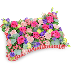 Shot from a slightly elevated oblique viewpoint on a clean white surface, this pillow-shaped floral tribute is a closely packed mosaic of colour and texture, designed to comfort as much as to commemorate. Hot fuchsia roses take center stage, their velvety faces catching the light with a soft glow, while clusters of pale pink carnations add a frilly, tactile counterpoint that invites a quiet touch. Interspersed among them are jewel-toned lisianthus in deep purple and sunny lemon-yellow ranunculus that punctuate the composition with playful brightness; tiny pink filler flowers add delicate, lace-like notes across the top. Astringent, silver-blue Eryngium thistles appear here and there, lending an architectural, slightly wild edge that contrasts with the softer blossoms. Underneath, generous swathes of green-rounded, silvered eucalyptus leaves and complementary foliage-create a cool, leafy bed whose scent evokes walks through Stanmore Common on an early morning. The pillow is finished with a ruffled, deep-red satin ribbon border that neatly frames and elevates the cushion shape, imparting elegance and a comforting formality appropriate for memorial settings. Prepared with the care you'd expect from Flowers Stanmore, the arrangement balances jubilant colour with respectful intention, making it suitable for funerals, commemorative services at Bentley Priory, or as a comforting sympathy gift delivered across Stanmore and nearby Harrow neighbourhoods. The composition is precise but gentle, the bouquet's overall effect a fragrant, visual embrace - a celebration of life rendered in lush petals, cool foliage, and considered texture.