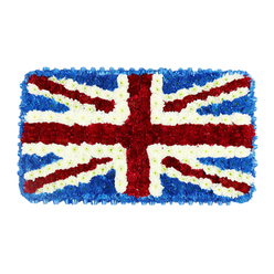 A beautifully composed floral artwork in a neat rectangular shape recreates the Union Jack flag with exceptional precision, resting against a simple white background that helps the colours appear vivid and uncluttered. The entire surface is formed from countless small blooms, placed so close together they form a continuous, cushion-like plane of petals. At the heart of the design, the broad vertical and horizontal arms of St. George's Cross are depicted with lush red flowers, likely compact chrysanthemums, their overlapping petals creating a thick, velvety stripe. From each corner of the rectangle, red diagonal bands emerge to represent St. Patrick's Cross, edged on both sides by slim lines of crisp white flowers that act as the fine fimbriation between red and blue. Wider swathes of white petals then stretch diagonally beneath these, forming St. Andrew's Cross and bringing a visual calm to the composition. The remaining spaces are filled with a dense carpet of rich blue blooms, possibly hydrangeas, forming a deep, textured backdrop and a tidy blue frame around the entire tribute. Gentle, diffused lighting reveals the natural softness and subtle curves of the petals, lending the flag a slightly raised, three-dimensional quality that differs from the flatness of fabric. This floral Union Jack feels reverent and considered, ideal for funerals, memorials, or civic ceremonies in and around Stanmore, where families might choose such a piece to honour military service, a lifetime of public duty, or simply a strong sense of British identity shared across generations in this quiet, leafy part of North West London.