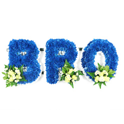 Captured against a stark white studio background, this three-part memorial spells BRO in bold, three-dimensional letters entirely covered with densely packed royal blue chrysanthemums. The blooms form a sumptuous, uniform surface that suggests both solidity and comfort; each tiny floret contributes to the plush silhouette of the B, R and O. At the lower curves and bases of the letters, delicate clusters of pale cream or soft yellow spray roses nestle with small white filler blooms-possibly mini chrysanthemums or gypsophila-balanced by an assortment of deep glossy leaves and lighter, lacy fern fronds. A thin white edging of the underlying foam or support is visible around the inner loops and perimeter, a reminder of the careful florist construction that keeps the composition neat and reverent. The combination of saturated blue and gentle ivory reads as peaceful and loyal, while the greenery grounds the piece with a live, earthy note. Under neutral studio lighting the textures are pronounced-velvety blue petals, the satin of rose buds, and the feathery movement of foliage-producing a quiet, consoling presence. This is an arrangement meant for memorial and funeral use, a bespoke tribute that speaks of kinship and memory. Locally, families might select this style for services at nearby venues or to place by a graveside at Stanmore's cemeteries or to send to a wake on Stanmore High Street; its respectful palette and disciplined craftsmanship make it an appropriate choice for those honouring a brother or dear friend.