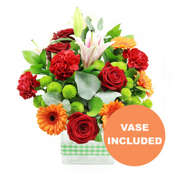 This exuberant mixed bouquet glows with cheerful colour, set against a clean white background that lets every detail shine, as though it were photographed in soft natural light drifting over Stanmore Hill. The arrangement is gathered in a modern, rectangular clear glass vase, the base wrapped in a light green and white gingham ribbon that lends a relaxed, homely charm. At the centre, deep red roses form the emotional core of the design, their tightly layered, velvety petals drawing the eye and conveying classic romance. Encircling them, bright orange gerbera daisies with deep brown centres open out like bright, optimistic faces, offering a strong contrast to the richer reds. Ruffled red-orange carnations are scattered through the bouquet, their frilly petals and rounded forms adding volume, movement, and a softer textural note. Above this vibrant cluster, several white oriental lilies stretch upward; some flowers are fully open, their trumpet-shaped petals marked with delicate pink veins and dusted stamens, while others remain in elegant, closed buds that promise ongoing interest. Between these larger focal blooms, small spheres of lime green pompom chrysanthemums nestle close to the stems, bringing a crisp, contemporary feel and a fresh jolt of colour that keeps the composition light and lively. The flowers are supported by an array of foliage, including broad, glossy dark green leaves and slender stems of silvery eucalyptus that curve gently outward, echoing the natural greenery of Canons Park. In the bottom right of the image, a circular orange graphic slightly overlaps the vase, with the words VASE INCLUDED in bold white capitals, clearly indicating that the stylish container forms part of the gift. The finished bouquet feels like a joyful, well-balanced celebration, ready for same-day delivery around Stanmore to mark birthdays, graduations, or housewarmings.