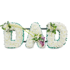 This photograph shows a dignified floral tribute spelling out "DAD" in upright, three-dimensional letters, designed for a funeral or memorial and displayed against a clean white background. Each letter is completely filled with tightly clustered white chrysanthemums, their small, button-like blooms forming a soft, uniform carpet of petals. The smooth, cushion-like texture of the flowers conveys calm, purity, and respect, with soft shadows falling between the petals to give a sense of depth. Around the edges, a rich emerald green satin ribbon has been carefully pleated and pinned in place, tracing the outline of each letter. The ribbon's subtle shine and deep colour create a striking contrast with the snowy white blooms, while faint hints of a dark support frame behind reveal that the tribute is constructed to stand securely at a service in Stanmore, perhaps in a local church or crematorium chapel.   

Gentle floral embellishments add personal touches to the otherwise uniform letters. On the top left curve of the first "D", a delicate spray of white freesia and lush green foliage softens the edge, suggesting a whisper of fragrance and natural movement. From this point, two slender green stems sweep diagonally across the design towards the centre, drawing the viewer's gaze into the "A". At their meeting point, near the lower part of the "A", an elegant asymmetrical focal arrangement appears: two deep purple calla lilies rise gracefully from a cluster of white freesia and greenery. Their sculpted, trumpet-shaped blooms and rich colour offer a quiet note of solemnity and depth, symbolising profound love and remembrance. On the final "D", at the upper right, a smaller cluster of white freesia and foliage echoes the first spray, restoring symmetry and flow across the tribute. The overall impression is of a serene, heartfelt farewell, a bespoke floral "DAD" arrangement that feels perfectly suited to honouring a father's life for families across Stanmore and the surrounding area.