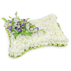 This rectangular floral pillow reads like a quiet benediction: hundreds of immaculate white chrysanthemum heads are arranged so tightly they form a continuous, velvety surface, evoking rest and purity. The gently rounded corners and slightly raised profile mimic the comforting shape of a sleeping cushion, while a light lime-green ribbon is pleated around the entire perimeter, creating a soft, scalloped border that brings a hint of renewal to the design. Crowning the upper-left corner is a modest but eloquent spray of blooms - white lisianthus with their soft, ruffled petals paired with deep purple lisianthus (several still in bud), introducing a note of solemn color and gentle contrast. Slender fern fronds and glossy, dark green foliage are woven through this cluster, lending texture and the faint green fragrance of freshly cut leaves; a darker green leaf wrap secures the stems and completes the tidy, respectful presentation. Set against a pristine white backdrop with a subtle drop shadow, the composition's tactile qualities-soft petals, lush greenery, and crisp ribbon-are clearly visible. The arrangement reads as an intimate expression of sympathy and remembrance, suitable for memorial services or a graveside tribute, and it communicates care through its meticulous craftsmanship and restrained palette. The overall impression is composed, consoling, and tender-a floral expression for those moments when comfort is needed more than words.