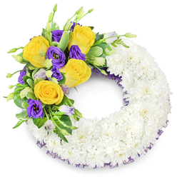 This circular sympathy wreath reads as an expression of calm: a generous base of small, immaculate white spray chrysanthemums forms a continuous ring, the petals so closely packed they create a soft, cloud-like cushion. The wreath's focal point occupies the upper-left curve, where golden yellow roses open fully, their layered petals suggesting warmth and friendship, and where deep purple lisianthus (Eustoma) add a velvety, somber contrast. A few unopened lisianthus buds in lighter lavender and pale green provide pauses of expectation and depth within the spray. Interwoven among the blooms is a selection of fresh green foliage - glossy rounded leaves, slender fern fronds and subtle variegated greenery - giving the composition body and a natural, garden-like resilience. Beneath the chrysanthemums, a fine dark purple crinoline or mesh peeks through at the inner and outer edges, a refined detail that quietly repeats the purple hues of the lisianthus and unifies the design. The lighting is neutral and clear, emphasizing textures: the softness of chrysanthemum petals, the satiny sheen of rose petals, the crispness of foliage. This arrangement is presented with the calm expertise of a Stanmore florist who has created tributes for local services near Stanmore Tube and Canons Park; it conveys respect, peaceful farewell, and an understated beauty appropriate for memorial settings without detracting from the solemnity of the occasion.