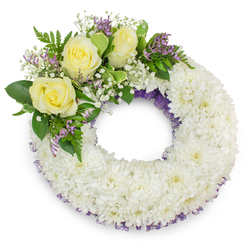 Presented against a stark white backdrop, this classic circular sympathy wreath is made predominantly of pristine white spray chrysanthemums, each tiny head packed close to form a cushion-like, almost clouded surface. The wreath keeps a perfect, solemn ring with an open centre that symbolizes unbroken love and continuity. A whisper of deep purple netting or ribbon is visible beneath the chrysanthemums on both the outer and inner rims, offering a refined, subtle contrast to the white. At the upper-left quadrant a composed cluster of three soft yellow roses provides a comforting focal point; their creamy petals are slightly cupped and scent the air faintly with a gentle, tea-rose sweetness. Around the roses, clouds of delicate white baby's breath and small sprays of pale lavender-purple limonium or statice add a lacy texture and a thoughtful hint of colour. Lush green foliage-broad leaves and feathery fern fronds-wraps the cluster and introduces a fresh, verdant backbone to the design. The arrangement is evenly lit with soft daylight tones, revealing the craftsmanship of each placement and casting a soft shadow that grounds the wreath on the white surface. As a local Stanmore florist might note when discussing memorial options for services near Stanmore Broadway or Bentley Priory, this wreath reads as both elegant and consoling, available in traditional sizes for delivery to funerals, wakes, or graveside services across the area.