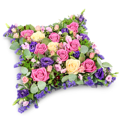 This image depicts a beautifully detailed floral cushion tribute, the kind often chosen for dignified farewell services in churches and crematoriums around Stanmore. The arrangement forms a soft-edged rectangle, like a plump pillow, positioned on a clean white background that allows every nuance of colour and texture to be clearly seen. The surface is entirely covered with fresh flowers, dominated by large roses in shades of vibrant fuchsia, gentle blush pink, and creamy pale yellow. Some roses are fully open, their spirals of petals unfolding in generous layers; others remain partially closed, creating a sense of depth and natural variation, as if the blooms are caught at different moments in time. Threaded between them are sumptuous deep purple lisianthus blooms, whose velvety, ruffled petals add drama and sophistication, much like the rich tones you might notice in established gardens near Stanmore Hill or Bentley Priory. Smaller blush pink spray roses are tucked in thoughtfully, their dainty heads bringing a touch of softness and innocence. Throughout the design, clusters of tiny lavender-blue filler flowers, likely statice or limonium, provide an airy, cloud-like effect, filling spaces and creating a gentle haze of cool-toned colour. Supporting this floral tapestry is a lush bed of foliage: rounded, silvery-green eucalyptus leaves introduce a cool, calming hue, while darker, fine-textured, fern-like greenery adds structure, shadow, and visual depth. The overall shape of the arrangement remains cushion-like and comforting, symbolising rest, peace, and the idea of cradling a loved one's memory. The mood is reflective and gentle, making this design a fitting floral tribute for funerals, memorial services, or graveside visits across the Stanmore area, where families seek a meaningful, beautifully composed expression of love and remembrance.