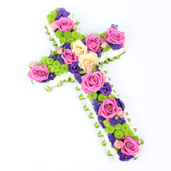 A beautifully crafted cross-shaped flower arrangement rests against a bright white background, angled gently so its form looks dynamic yet serene. The entire outline is traced with a generous border of white spray chrysanthemums, their tiny, cushiony blooms packed tightly to form a soft, comforting frame. Just beyond this, a slim edging of folded pale green ribbon or foliage runs around the cross, adding both definition and a touch of fresh, leafy colour that sets off the whiteness of the surrounding petals. Within this border, the central area of the cross is alive with richly coloured blooms. Large roses in hot fuchsia pink and soft creamy yellow form the main focal points, their open, velvety petals inviting the eye to linger. Between and around them, clusters of lime-green button chrysanthemums add structure and a lively, textural rhythm, their spherical forms lifting the composition visually. Delicate flowers in deep purple and violet-resembling lisianthus or statice-are woven through the spaces, providing contrast and a sense of depth that gives the tribute dignity and emotional weight. The lighting is clear and bright, ensuring each flower type, from the smallest white floret to the boldest rose, can be distinguished. Created with the kind of care you'd expect from an experienced Stanmore florist, this floral cross would be a fitting choice for a funeral, wake, or memorial at a local church near Stanmore Hill, expressing sympathy, spiritual comfort, and a quiet celebration of the memories shared with the person being honoured.