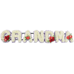 The image shows a tender and elegant floral tribute spelling out "GRANDMA" in large capital letters, carefully arranged in a straight line against a simple white background so that every detail of the design can be clearly appreciated. Each letter is formed entirely from densely packed white cushion chrysanthemums, the neat, domed flowers arranged so closely that they create a soft, fluffy texture, like a continuous white blanket of petals. Around the edges, a trim of delicate pale lilac ribbon subtly outlines each character, offering a gentle purple sheen that adds refinement and echoes the quiet calm of Stanmore's tree-lined streets. Vivid red highlights are placed thoughtfully throughout the tribute. At the beginning, the letter "G" is adorned with a compact cluster of deep red roses and a distinctive red-and-white variegated calla lily, its sleek, funnel-shaped bloom curling elegantly and lending a modern twist. The first "A" features a single, heart-shaped red anthurium lily at its base, the polished surface and central spadix giving a sculptural contrast to the feathery chrysanthemums. In the middle of the word, the letter "M" is embellished with another group of red roses paired with a second red anthurium, drawing the eye to the centre as a symbol of the grandmother's central place in family life. The final "A" finishes the sequence with one more red anthurium bloom, creating balance from start to finish. The letters sit on a discreet dark green structural stand that lifts them slightly, providing support without distracting from the flowers. Crafted with care, this personalised funeral arrangement would sit beautifully in a Stanmore chapel or cemetery, offering a heartfelt farewell infused with quiet dignity and enduring love.