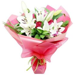 A luxurious floral arrangement of white lilies and deep red roses is shown against a bright white backdrop, every petal, leaf, and fold of wrapping clearly defined. The lilies, with their large, open trumpets and gently recurved petals, take centre stage, their pure white surfaces acting like soft reflectors of light. In the middle of each bloom, dark, chocolate-brown anthers sit atop slender filaments, their colour echoing the richness of the red roses below. Several lily buds, tall and slender with green outer casings, rise from the bouquet, promising continued blooms once the flowers are delivered to a home in Stanmore. Interwoven with the lilies, the red roses provide a velvety contrast, some buds tightly closed, others beginning to loosen into layered spirals that convey romance and devotion. Around and behind the blooms, abundant greenery in deep and mid-green tones fills the spaces, with broad, glossy leaves catching subtle highlights and fine, grass-like stems adding vertical lines and texture, reminiscent of the gentle sway of trees around Bentley Priory Nature Reserve. The whole composition is wrapped generously in shiny, bright pink cellophane that appears slightly translucent, its crinkled, ruched folds forming a flared collar around the flowers. At the point where the wrap narrows to hold the stems, a narrow lime green satin ribbon is tied, its crisp bow adding a playful yet refined touch. This bouquet feels both classic and contemporary, ideal for celebrating milestones, saying "thank you," or sending love within Stanmore, and reflects the skill and attention to detail you would expect from a dedicated neighbourhood florist.