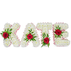An elegant, custom-made name tribute spells KATE in bold, three-dimensional block letters, each letter painstakingly formed from a thick carpet of small white chrysanthemums that give the design a soft, cloud-like volume. The petals are layered close together so the letters read pure and luminous beneath a calm, neutral background. Each letter is edged with a slim pale pink satin ribbon that creates a refined border and a gentle visual warmth against the crisp white. A single deep red rose is set into the centre of every letter, its velvety petals offering a striking focal point framed by a generous spray of green foliage-broad glossy leaves and fine fern fronds adding movement and depth. On the 'K' the rose nestles on the lower portion of the left vertical stroke; the 'A' bears its rose at the peak of its triangular top; the 'T' is punctuated with a rose at the base of the left side of the horizontal bar; and the 'E' carries its floral accent on the middle horizontal arm. The combined palette of white chrysanthemums, soft pink ribbon, vivid red roses, and rich greens reads as timeless and respectful, ideal for funeral services and memorial displays. The subtle scent of roses against the cool chrysanthemum base and the tactile ribbon edging convey careful craftsmanship. As Stanmore florists, Flowers Stanmore often prepare such personalised tributes for families attending services near Stanmore Tube, Canons Park, or small chapels off the High Street, and we can advise on delivery and placement for local memorials.