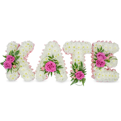 An expert-crafted floral tribute spells the name 'KATE' in bold, three-dimensional letters, each formed from a tightly packed carpet of small, pristine white chrysanthemums that produce a seamless, cloud-like texture. A soft, pale-pink satin ribbon traces the inner contours of every character, lending a refined, blush-edged silhouette to each letter and enhancing the gentle feminine mood. Strategic clusters of hot pink roses punctuate the white field: the K is accented with two fully opened pink roses on its left side set among mixed green leaves; the A bears a single, prominent rose toward its base framed by curling sprigs and tiny pink buds; the T is anchored by one vivid rose at its bottom center with delicate fern-like foliage; and the E carries a pair of roses along its right edge with variegated greens and soft pink accents. The scent is quietly rose-led, layered beneath the clean, slightly green perfume of the chrysanthemums and foliage. Presented as a freestanding display on a pristine white surface, subtle under-shadows reveal the arrangement's depth and careful wiring. This kind of bespoke lettering is often chosen for funerals or memorial services, or as a deeply personal celebration of life. As Flowers Stanmore, we prepare these pieces with local sensitivity - imagining this tribute delivered to a chapel near Stanmore Hill or set beside floral tributes at Stanmore Broadway - offering a dignified and personal expression of love and remembrance.