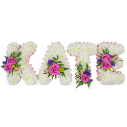 In this carefully composed memorial piece, the name KATE stands letter by letter in elegant, upright forms constructed from thousands of white chrysanthemum heads, each flower pressed close to its neighbour to create a smooth, pillowy surface and a strong architectural silhouette. A subtle satin ribbon in a pale pink hue neatly borders the letters, picking out the shapes and lending a soft, ribboned glow that reads as gentle and reverent under the neutral lighting. Vibrant accent clusters punctuate the white field: large fuchsia-pink roses anchor each spray, accompanied by smaller pastel pink spray roses for softness, and interlaced with deep purple or blue statice or lisianthus for contrast and small, textured accents. The arrangement is intentionally placed: a prominent grouping sits on the diagonal of the K, a bloom cluster rests by the lower right of the A, the T bears a compact spray at its foot, and the E carries two well-balanced clusters along the central and lower bars. Varied greenery-delicate fern fronds, longer eucalyptus leaves, and subtle filler foliage-frames each bouquet, creating depth, movement, and a gentle herbal scent that complements the roses. The visual language is comforting: crisp white signals purity and remembrance while the pinks and purples convey affection and solemnity. Created with the quiet care of a local Stanmore floral designer, this bespoke tribute suits funerals, wakes, or celebration-of-life ceremonies held near Canons Park, Stanmore High Street, or the surrounding community, offering a personalised and heartfelt way to honour someone called Kate. Flowers Stanmore prepared this design with attentive craftsmanship and a focus on proportion and sentiment.
