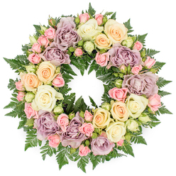 A beautifully detailed circular wreath is shown against a clean white backdrop, making every rose and fern frond clearly visible. The arrangement is dense and luxurious, with no bare spaces, giving the impression of a continuous, comforting embrace. Dominating the design are large, open roses in soft ivory and creamy white, their many petals layered like fine silk and curling slightly at the tips. Nestled among them are clusters of small blush pink spray roses, along with tiny buds that are just beginning to open, bringing a sense of delicacy and unfolding life. Gentle accents of peach and buttery yellow roses warm the overall palette, while distinctive dusty lavender and muted mauve blooms add a cooler, vintage note that feels both refined and soothing. The flowers are supported by a rich backdrop of dark green fern foliage and other finely textured greenery, whose feathery fronds weave between the petals, adding depth, contrast, and a fresh, garden-like feel. The circle itself is perfectly formed, a traditional symbol of unending love and remembrance. The mood is calm and reflective, ideal for use as a funeral wreath or memorial tribute in Stanmore, perhaps at a service near Canons Park or along the quieter residential streets that give the area its village atmosphere. Carefully arranged by experienced local florists, this wreath provides a gentle, dignified way to express sympathy, respect, and enduring affection during times of loss.