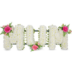 The image portrays a heartfelt floral tribute spelling "MOM," carefully photographed against an uncluttered white background so that every detail stands out. Three large capital letters sit side by side, each one built on a sturdy frame and completely blanketed in white chrysanthemums. These are small, spray-type chrysanthemums, packed tightly together so their rounded blooms merge into a soft, even surface that resembles a cushion or a snowy hillside. The pure white petals convey innocence, remembrance, and peace, lending the tribute a quiet dignity that suits a solemn farewell. Along the lower edge of the letters, a slim band of dark green-likely a ribbon over a metal frame-adds a subtle outline and hints at the solid construction beneath the sea of petals. Breaking the expanse of white, three complementary clusters of pink flowers and greenery introduce gentle colour. At the top left of the first "M," a standout fuchsia rose sits at the centre, surrounded by paler pink spray roses and rosebuds, with delicate fern fronds adding a lacy, feathered texture. A similar grouping ornaments the lower area between the "O" and the second "M," while another blooms at the top right of the final "M," balancing the composition visually. The interplay of bright white and vivid pink creates a sense of warmth within the stillness, evoking gratitude and enduring affection for a beloved mother. This kind of "MOM" tribute would feel at home in a Stanmore chapel, on a hearse travelling down Marsh Lane, or placed lovingly by a graveside, a soft-spoken yet unmistakable declaration of love expressed through flowers.