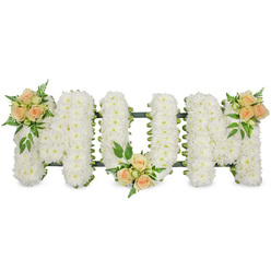 The photograph shows a lovingly designed sympathy arrangement spelling "MOM" in large, upright block letters, set against a crisp white background that keeps all focus on the flowers. Each letter is completely covered with pure white flowers, almost certainly small spray chrysanthemums, arranged so tightly that they form a smooth, continuous surface with no gaps. The countless little petals create a soft, velvety texture that appears calm and comforting, ideal for a funeral service or celebration of life around Stanmore and the surrounding areas. A neat dark green framework and ribbon edge subtly outline each letter, giving the word strength and clarity while hinting at the skilled construction beneath. Adding warmth and personality, three graceful clusters of spray roses in soft peach and apricot tones are placed along the tribute. One cluster rests on the upper left section of the first "M", another nestles at the lower central point of the "O", and the third decorates the upper right of the final "M", creating a gentle rhythm of colour from left to right. The roses are a mix of buds and partially open blooms, their curved petals catching the light, and each grouping is surrounded by feathery ferns and glossy green leaves for contrast and natural texture. The piece feels both elegant and heartfelt, reflecting the love of children and family members wishing to honour a mother's life in Stanmore, perhaps after a service near the high street or close to Bentley Priory nature reserve. The contrast between the serene white base and the warm peach accents symbolises both peace and enduring affection, making this "MOM" letter tribute a quietly powerful expression of farewell and remembrance.